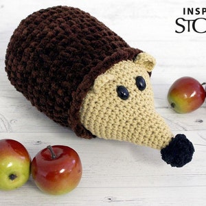 Puede incluir: Un juguete de erizo de punto de crochet hecho con hilo marr&oacute;n y beige, ojos negros y nariz negra. El erizo est&aacute; sentado sobre una superficie de madera blanca con tres manzanas rojas.