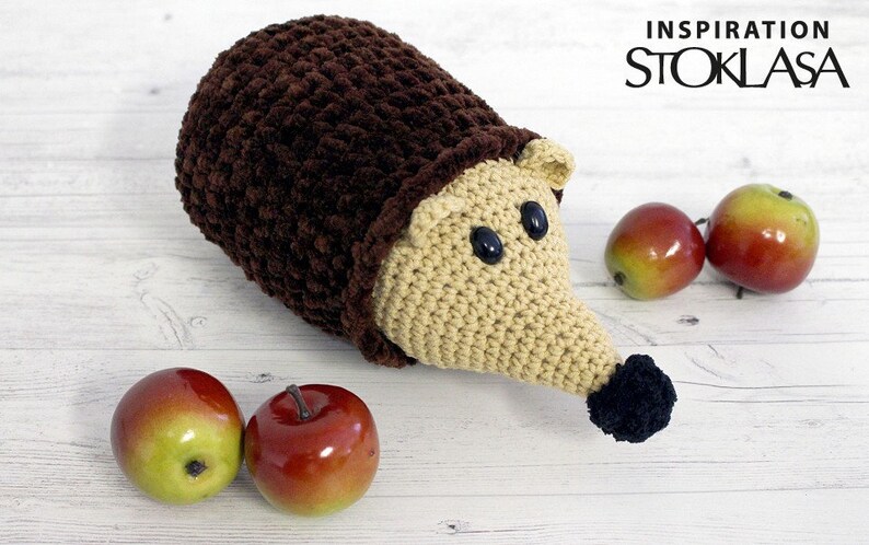 Puede incluir: Un juguete de erizo de punto de crochet hecho con hilo marr&oacute;n y beige, ojos negros y nariz negra. El erizo est&aacute; sentado sobre una superficie de madera blanca con tres manzanas rojas.
