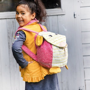 May include: A young girl wearing a yellow vest with a pink and white animal-shaped backpack. The backpack has a pink polka dot pattern and a white stripe pattern. The backpack has a fluffy white face with black eyes and a pink nose.
