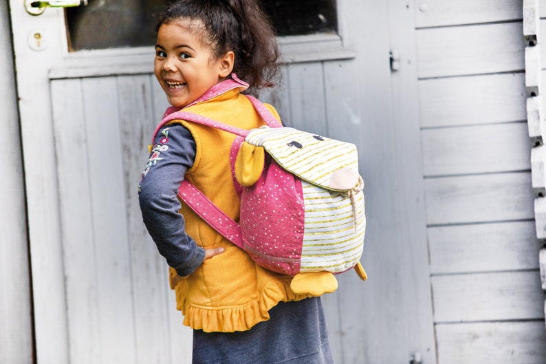 May include: A young girl wearing a yellow vest with a pink and white animal-shaped backpack. The backpack has a pink polka dot pattern and a white stripe pattern. The backpack has a fluffy white face with black eyes and a pink nose.