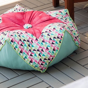 Puede incluir: Un coj&iacute;n de suelo grande, redondo y multicolor con una parte superior acolchada de color rosa. El coj&iacute;n est&aacute; hecho de tela con un patr&oacute;n geom&eacute;trico de tri&aacute;ngulos en tonos de rosa, verde, blanco y negro. El coj&iacute;n est&aacute; sentado en un suelo de baldosas grises.