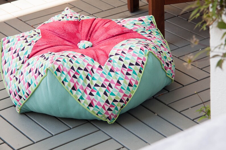 Puede incluir: Un coj&iacute;n de suelo grande, redondo y multicolor con una parte superior acolchada de color rosa. El coj&iacute;n est&aacute; hecho de tela con un patr&oacute;n geom&eacute;trico de tri&aacute;ngulos en tonos de rosa, verde, blanco y negro. El coj&iacute;n est&aacute; sentado en un suelo de baldosas grises.