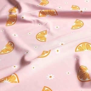 Op de afbeelding: Roze stof met een herhalend patroon van oranje citrusvruchtenplakjes en witte madeliefjes.