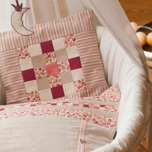 May include: A white wicker bassinet with a pink and white patchwork pillow. The pillow has a floral pattern and a red and white striped border.