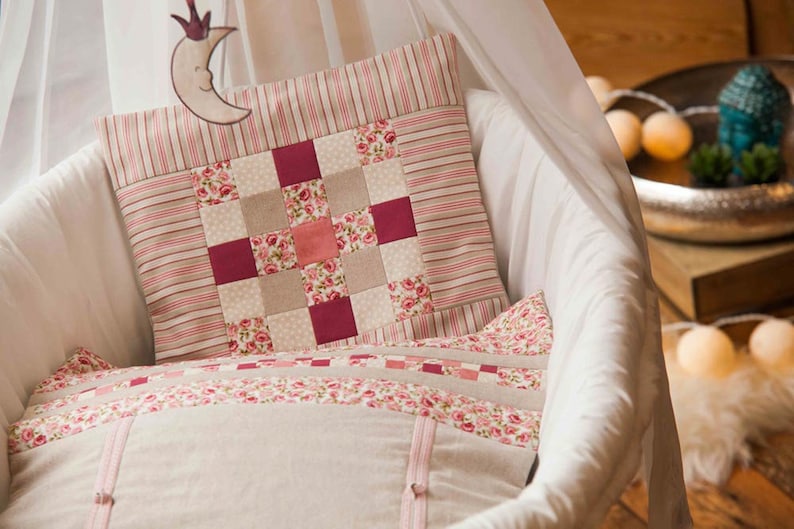 May include: A white wicker bassinet with a pink and white patchwork pillow. The pillow has a floral pattern and a red and white striped border.