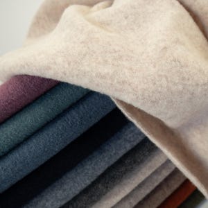Könnte beinhalten: Ein Stapel verschiedenfarbiger Wollstoffmuster. Die oberste Schicht ist hellbeige, darunter liegen Schichten aus lila, teal, blau, grau und braun. Die Stoffe haben ein weiches, strukturiertes Aussehen.