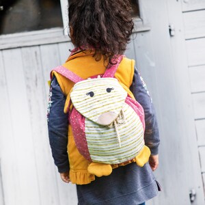 May include: A child wearing a blue and yellow jacket with a pink and white animal-shaped backpack. The backpack has a white and gold striped front with a pink nose and black eyes.