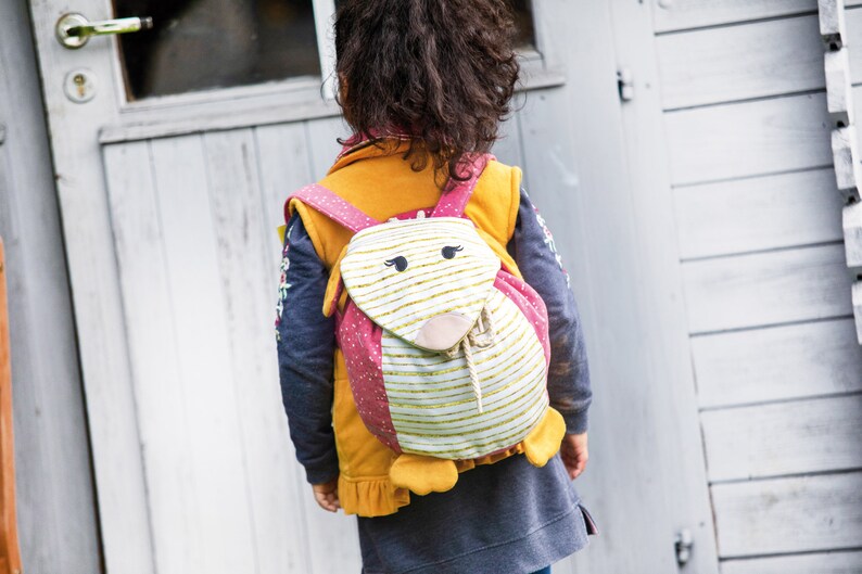 May include: A child wearing a blue and yellow jacket with a pink and white animal-shaped backpack. The backpack has a white and gold striped front with a pink nose and black eyes.