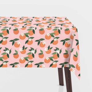 Könnte beinhalten: Eine rosa Tischdecke mit einem sich wiederholenden Muster aus Orangenscheiben und weißen Blüten. Die Tischdecke ist über einen Tisch mit braunen Beinen gelegt.