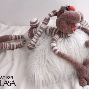 Puede incluir: Un juguete de conejo de crochet a rayas marrones y blancas con extremidades largas y una nariz roja. El conejo est&aacute; acostado sobre una almohada blanca y esponjosa.