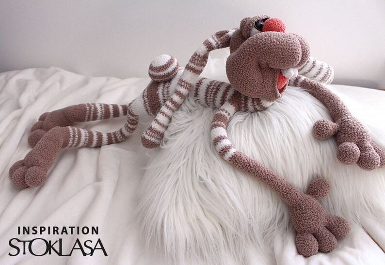 Puede incluir: Un juguete de conejo de crochet a rayas marrones y blancas con extremidades largas y una nariz roja. El conejo est&aacute; acostado sobre una almohada blanca y esponjosa.