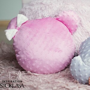 Puede incluir: Una almohada de felpa rosa con un lazo blanco y una superficie texturizada. La almohada tiene forma de cabeza de oso con dos orejas peque&ntilde;as.