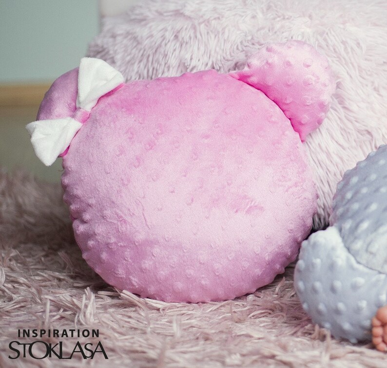 Puede incluir: Una almohada de felpa rosa con un lazo blanco y una superficie texturizada. La almohada tiene forma de cabeza de oso con dos orejas peque&ntilde;as.