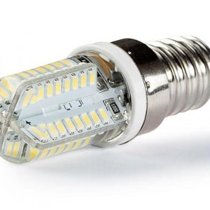 Könnte beinhalten: Eine kleine, silberne LED-Glühbirne mit einem klaren Glasgehäuse. Die Glühbirne hat einen Standard-Schraubsockel und verfügt über mehrere kleine LEDs, die in einem Kreis angeordnet sind.