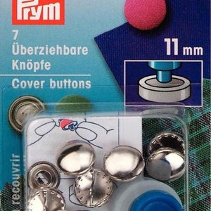 Op de afbeelding: Een verpakking met 7 zilveren knopen met een diameter van 11 mm, met een blauw plastic gereedschap om de knopen te bevestigen. De verpakking is gelabeld met "Prym" en "Cover buttons".