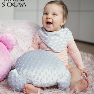 Puede incluir: Un beb&eacute; con un mono rosa de manga larga con un babero de estampado de nubes grises y blancas, sentado en una alfombra rosa peluda con una almohada de felpa gris. El beb&eacute; sonr&iacute;e y mira hacia arriba.