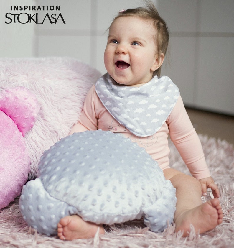 Puede incluir: Un beb&eacute; con un mono rosa de manga larga con un babero de estampado de nubes grises y blancas, sentado en una alfombra rosa peluda con una almohada de felpa gris. El beb&eacute; sonr&iacute;e y mira hacia arriba.