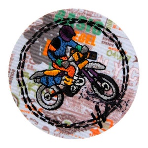 Op de afbeelding: Geborduurd embleem met een motorcrosser op een crossmotor. Het embleem is rond en heeft een zwart gestikte rand. De rijder draagt blauwe, oranje en paarse kleding. De achtergrond heeft een patroon met groene en oranje tekst.