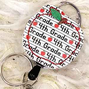 May include: A red apple-shaped retractable badge holder with a white background and black text that reads "4th Grade". The apple has a green leaf and a red stem.