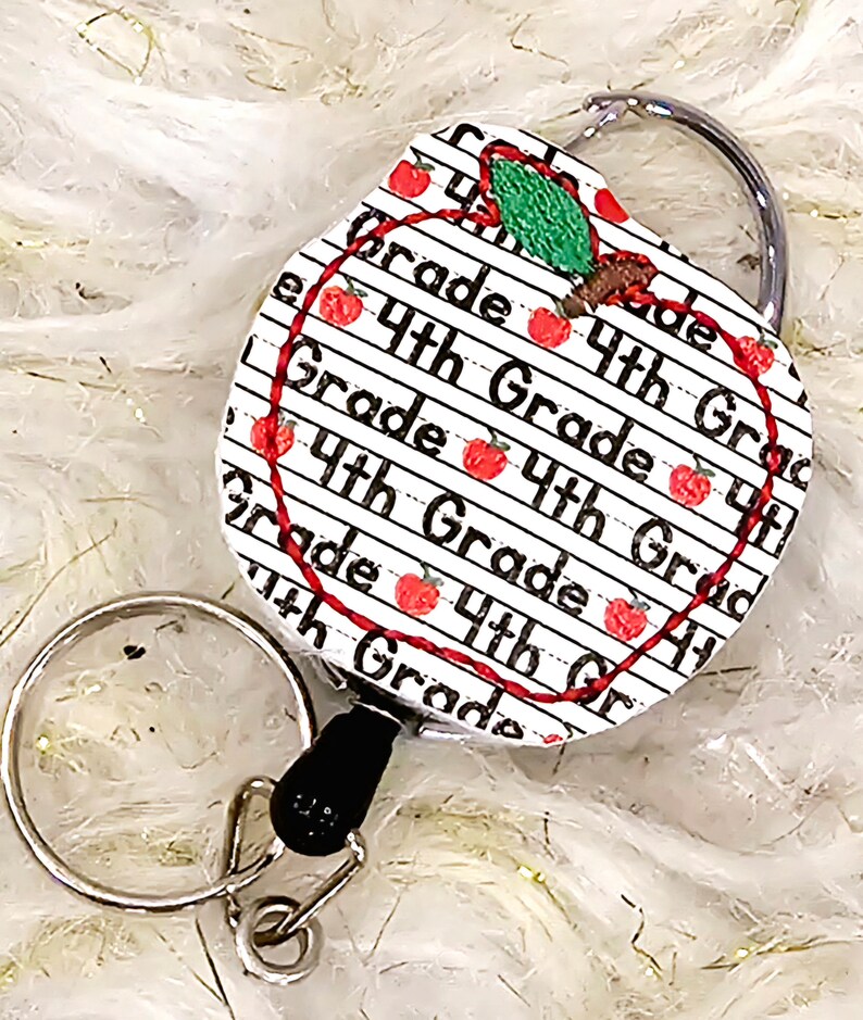 May include: A red apple-shaped retractable badge holder with a white background and black text that reads "4th Grade". The apple has a green leaf and a red stem.