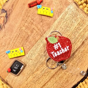May include: A red glitter apple with a green stem and the text "#1 Teacher" on it. The apple is attached to a silver retractable badge holder with a clear strap.