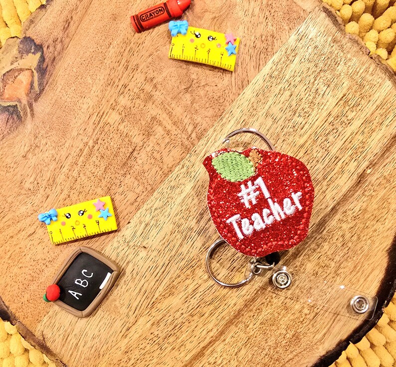 May include: A red glitter apple with a green stem and the text "#1 Teacher" on it. The apple is attached to a silver retractable badge holder with a clear strap.