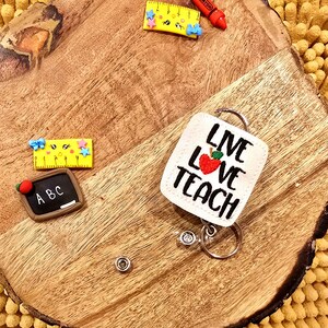 May include: White fabric badge holder with the text "Live Love Teach" and a red apple with a green leaf. The badge holder is attached to a silver retractable badge reel.