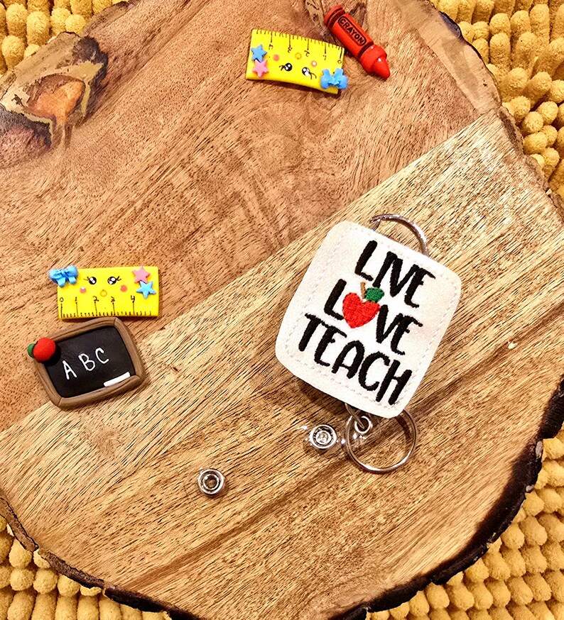May include: White fabric badge holder with the text "Live Love Teach" and a red apple with a green leaf. The badge holder is attached to a silver retractable badge reel.