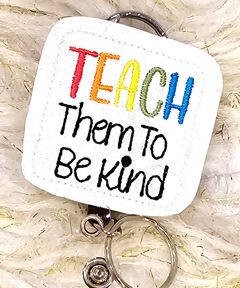 May include: White square badge holder with a rainbow colored "TEACH" and the text "Them to Be Kind" in black. The badge holder is attached to a silver swivel clip.