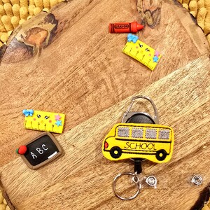 May include: A yellow school bus keychain with a retractable badge holder. The keychain features a black and white embroidered school bus with the word "SCHOOL" on the side. The keychain also includes a silver swivel clip, a silver key ring, and two silver badge holders.