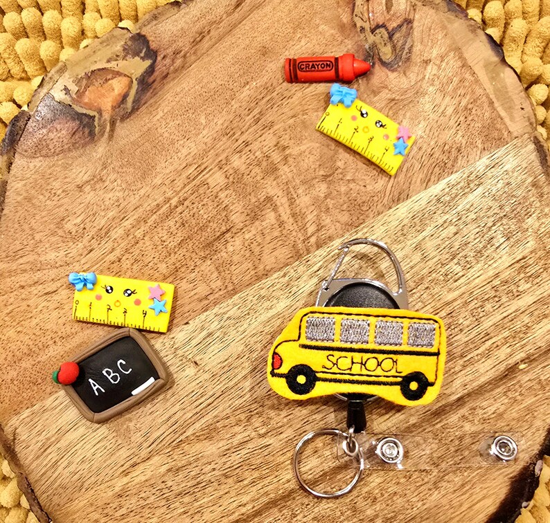 May include: A yellow school bus keychain with a retractable badge holder. The keychain features a black and white embroidered school bus with the word "SCHOOL" on the side. The keychain also includes a silver swivel clip, a silver key ring, and two silver badge holders.