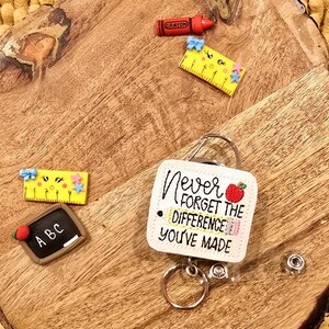 May include: White square badge holder with a red apple and a yellow pencil, with the text "Never forget the difference you've made". The badge holder is attached to a retractable badge reel with a silver key ring.