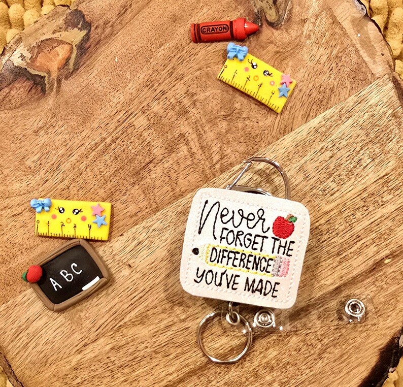 May include: White square badge holder with a red apple and a yellow pencil, with the text "Never forget the difference you've made". The badge holder is attached to a retractable badge reel with a silver key ring.