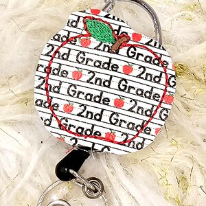 May include: A white apple-shaped retractable badge holder with a red outline and a green leaf. The apple is covered in a pattern of red apples and the text "2nd Grade" repeated multiple times.