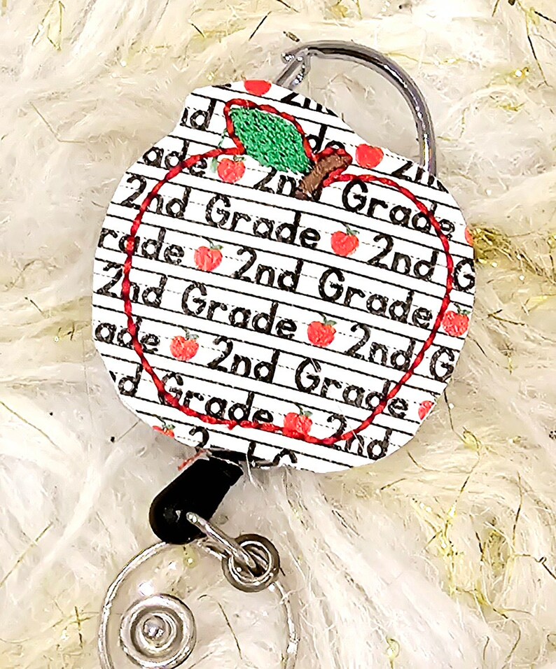 May include: A white apple-shaped retractable badge holder with a red outline and a green leaf. The apple is covered in a pattern of red apples and the text "2nd Grade" repeated multiple times.