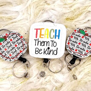 May include: Three white fabric badge holders with retractable reels. One holder features a red apple with the text "2nd Grade" repeated around it. Another holder features a red apple with the text "4th Grade" repeated around it. The third holder has the text "TEACH Them To Be Kind" in rainbow colors.