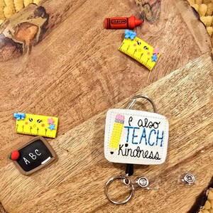 May include: A white badge holder with a yellow pencil and the text "I also teach kindness". The badge holder is attached to a retractable badge reel with a silver key ring. There are also several small school-themed decorations on a wooden surface.