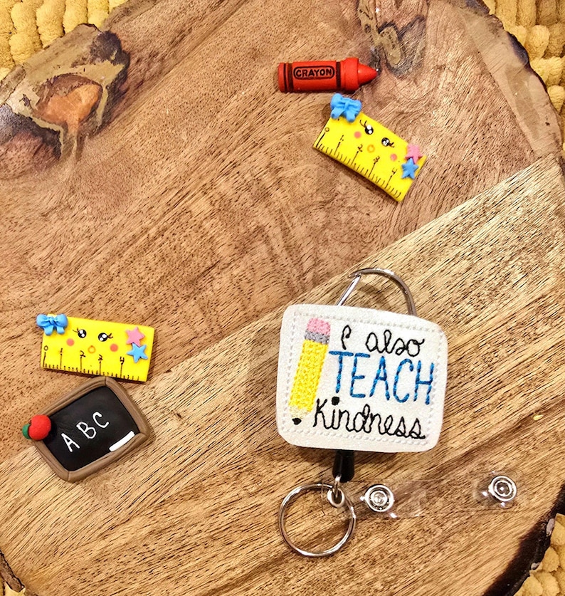 May include: A white badge holder with a yellow pencil and the text "I also teach kindness". The badge holder is attached to a retractable badge reel with a silver key ring. There are also several small school-themed decorations on a wooden surface.