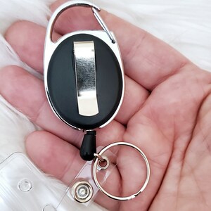 May include: A silver retractable badge reel with a black plastic clip and a clear plastic badge holder. The reel has a silver swivel hook and a silver ring.