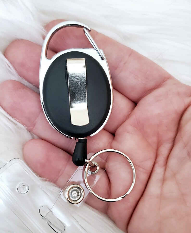 May include: A silver retractable badge reel with a black plastic clip and a clear plastic badge holder. The reel has a silver swivel hook and a silver ring.