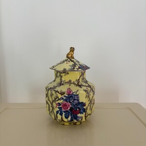May include: A yellow ceramic jar with a lid, featuring a floral design with pink and blue roses. The jar has a gold-colored cat figurine on top of the lid. The jar is decorated with a vine pattern.