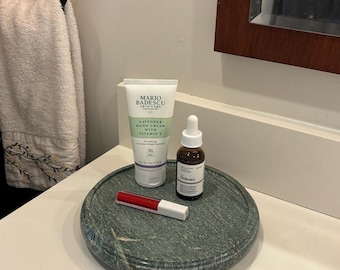 Puede incluir: Una bandeja de mármol verde con productos para el cuidado de la piel: un tubo de crema de manos de lavanda Mario Badescu, un frasco de suero The Ordinary y un brillo labial rojo. La bandeja es redonda.
