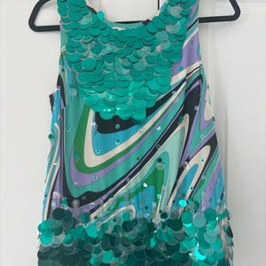 Puede incluir: Una blusa sin mangas con un vibrante estampado en espiral en tonos turquesa, morado, verde y blanco. La blusa está adornada con lentejuelas turquesas grandes alrededor del cuello y el dobladillo. La prenda cuelga de una percha negra.