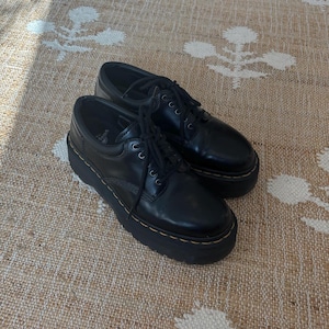 Può includere: Un paio di scarpe platform in pelle nera con lacci neri. Le scarpe hanno una suola spessa nera con cuciture gialle sul perimetro. Le scarpe sono su un tappeto beige e bianco fantasia.