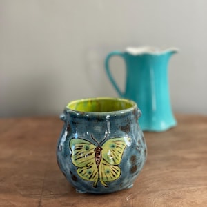May include: A handmade ceramic pot with a blue exterior and a yellow butterfly design. The interior is bright yellow. A turquoise pitcher is in the background. The pot has small handles and sits on a wooden surface.