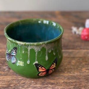 May include: A green ceramic bowl with a teal interior and a glossy finish. The bowl features two butterfly decals with black, orange, and blue wings. The rim has a white, dripped glaze effect, adding a unique touch to the handmade pottery.