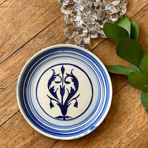 May include: A ceramic plate with a cream-colored center and a blue floral design. The rim features concentric blue stripes. The plate is on a wooden surface, with a crystal chandelier and green leaves in the background.