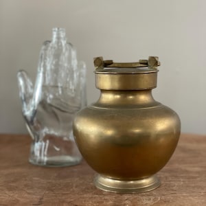 May include: A vintage brass container with a lid and a clear glass hand-shaped bottle. The brass container has a rounded body and a pedestal base. The glass bottle is in the background.