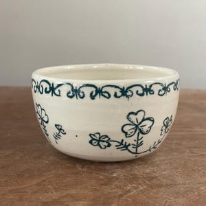 May include: A small, off-white ceramic bowl with a dark teal floral design. The bowl features a decorative border and stylized flowers around the exterior. The bowl is set on a wooden surface.
