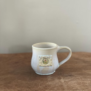 May include: A cream-colored ceramic mug with a curved handle. The mug features a small, rectangular image with gold accents. The image appears to be a painting or illustration. The mug is on a brown surface.